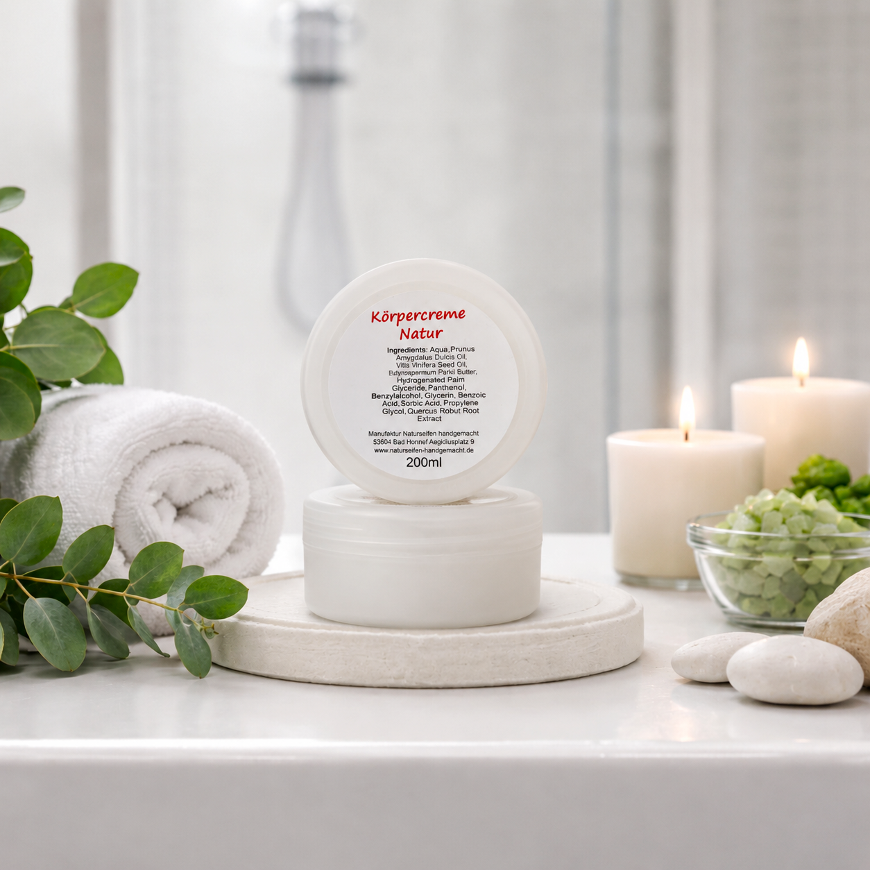 Im Vordergrund weiße Runddose Körpercreme Natur, stehend auf Waschbeckenanrichte mit Kerzen und Dekos